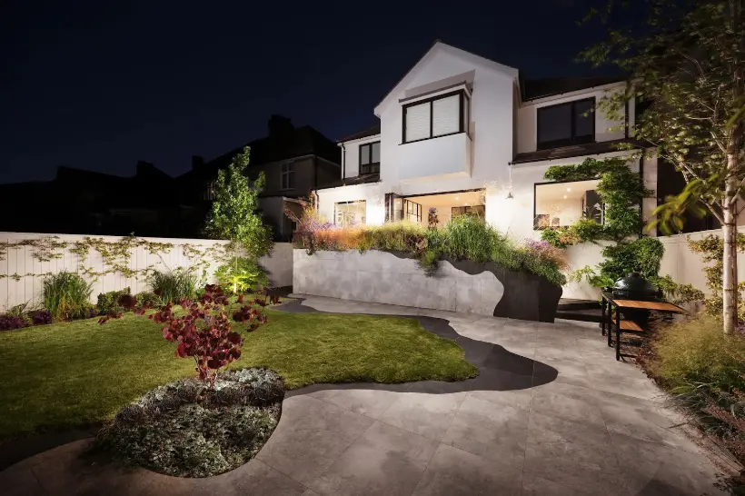 a garden at night, showing a tidy garden design looping up to a house via a staircase covered in greenery